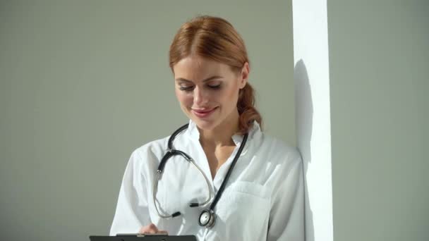 Una joven doctora caucásica vestida con un abrigo médico blanco y un estetoscopio escribe una historia clínica en un cuaderno. Médico sonriente posando en la oficina del hospital — Vídeos de Stock