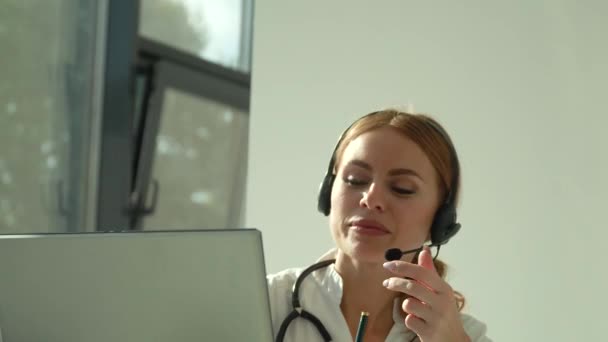 Professionell kvinnlig läkare i vit medicinsk rock och headset gör konferens samtal på bärbar dator, konsultation distans patient online i videochatt — Stockvideo