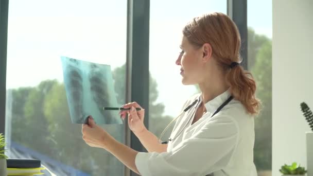 Ärztin betrachtet Röntgenbild der Lungen, während sie in moderner Klinik am Tisch sitzt — Stockvideo