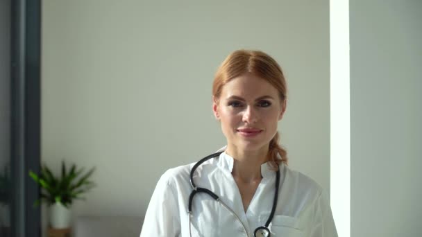 Mulher branca jovem médico vestindo casaco médico branco olhando para a câmera e mostra pílulas. Médica sorridente posando no consultório do hospital — Vídeo de Stock