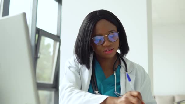 Aantrekkelijke Afrikaanse Amerikaanse vrouwelijke arts met behulp van laptop aan het bureau tijdens het werken in het medisch kantoor — Stockvideo