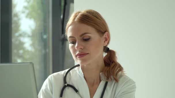 Attractive caucasian female doctor in white coat and stethoscope over her neck using laptop at desk, looking at camera while working in medical office — Stock Video