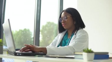 Beyaz önlüklü çekici Afrikalı kadın doktor ve boynunda steteskop. Tıp bürosunda çalışırken bilgisayarını kullanıyor.