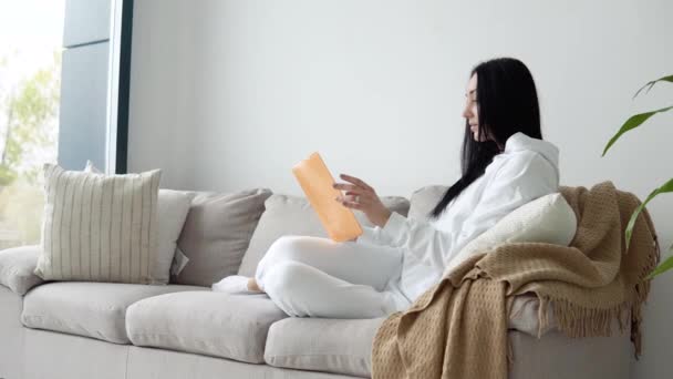 Mujer joven leyendo un libro mientras está sentada en un sofá en una sala de estar, leyendo libros. Quédate en casa concepto — Vídeos de Stock