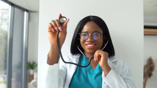 Αφροαμερικανή γιατρός με λευκό παλτό σε κλινική στο χώρο εργασίας κρατά στηθοσκόπιο στο χέρι του και ακούει την αναπνοή του πιθανού ασθενούς. Γυναίκα γιατρός χρησιμοποιεί στηθοσκόπιο — Αρχείο Βίντεο