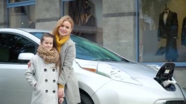 Kızıyla birlikte bir kadın elektrikli arabasının yanında duruyor ve otoparktaki kameraya bakıyor. Benzin istasyonunda elektrikli araba şarj etmek.