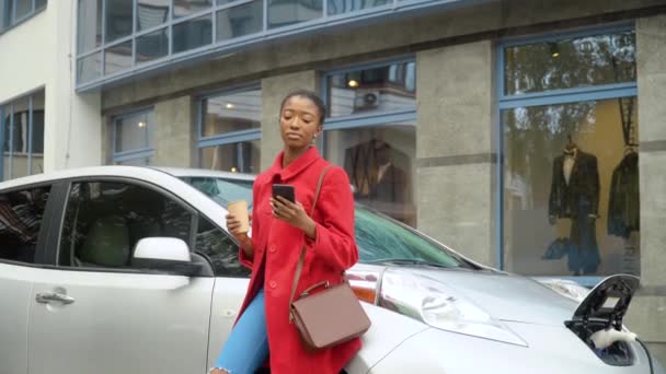 Afroamerykanka używa smartfona i opiera się na elektrycznym samochodzie, który się ładuje. Transport przyjazny dla środowiska — Wideo stockowe