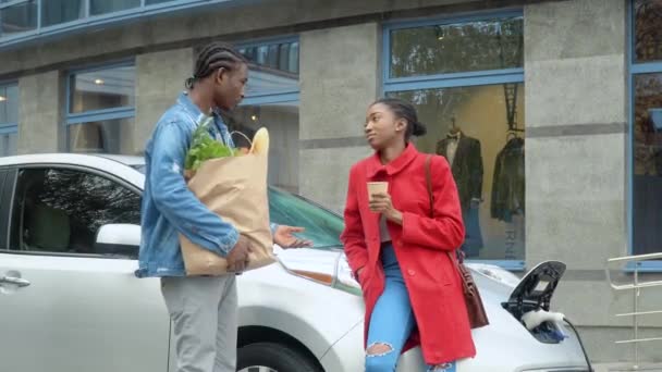 Afro-Amerikaans echtpaar staat met kruidenierswaren in de buurt van elektrische auto. Elektrische auto opladen bij het elektrische benzinestation — Stockvideo