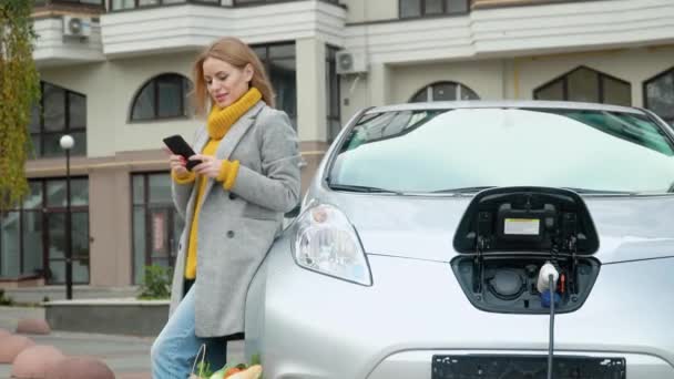 スマートフォンを使用している女性と充電している電気自動車にもたれます。電気自動車を充電するプラグ。電気自動車の充電。環境に優しい交通機関 — ストック動画