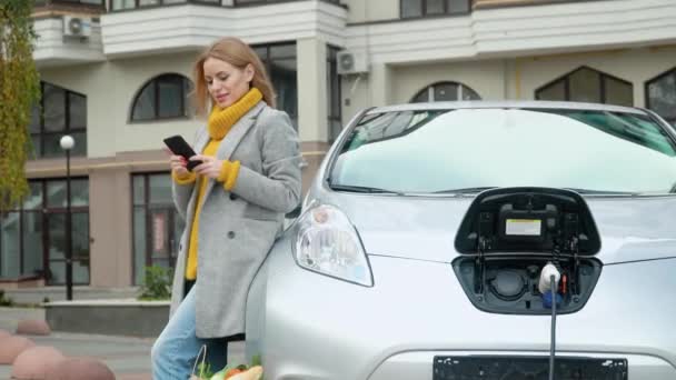 Donna che utilizza smartphone e si appoggia su auto elettrica che si sta caricando. Spina di ricarica di un'auto elettrica. Ricarica auto elettrica. Trasporti ecologici — Video Stock