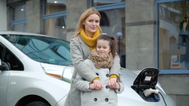 Egy nő a lányával az elektromos autója mellett áll és a kamerába néz. Feltölteni egy elektromos autót egy benzinkútnál. Környezetbarát szállítás — Stock videók