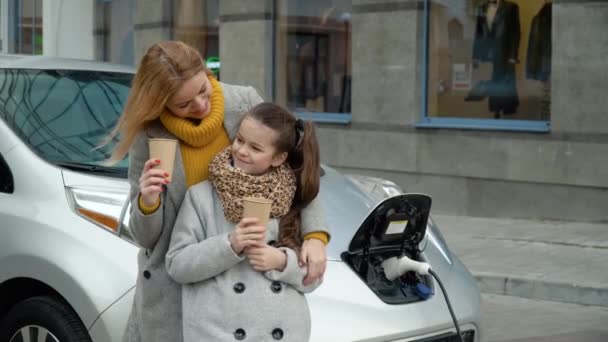 娘と一緒に電気自動車の近くでコーヒーを飲む女性。ガソリンスタンドでの電気自動車の充電 — ストック動画