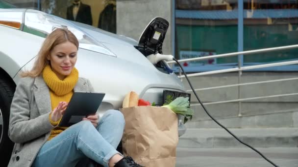 Vrouw gebruik tablet en leunt op elektrische auto die wordt opgeladen. Plug laadt een elektrische auto op. Opladen van elektrische auto — Stockvideo