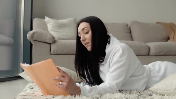 Молодая красивая женщина читает книгу, лежащую на полу дома — стоковое видео
