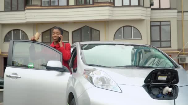 Αφροαμερικανή να μιλάει στο τηλέφωνο ενώ φορτίζει ένα ηλεκτρικό αυτοκίνητο. Φιλικές προς το περιβάλλον μεταφορές — Αρχείο Βίντεο