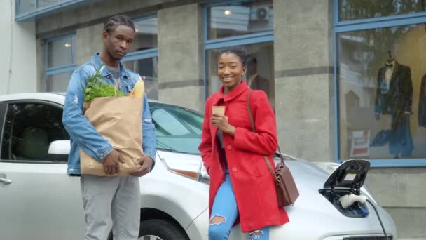 Afro-Amerikalı çift, markette elektrikli arabanın yanında durup kameraya bakıyorlar. Elektrikli benzin istasyonunda elektrikli araba şarj ediliyor. — Stok video