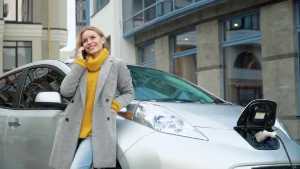 女性は電気自動車を充電しながら電話で話しています。環境に優しい交通機関 — ストック動画
