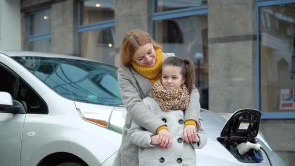 Kobieta z córką stoi blisko elektrycznego samochodu i patrzy w kamerę. Ładowanie elektrycznego samochodu na stacji benzynowej — Wideo stockowe