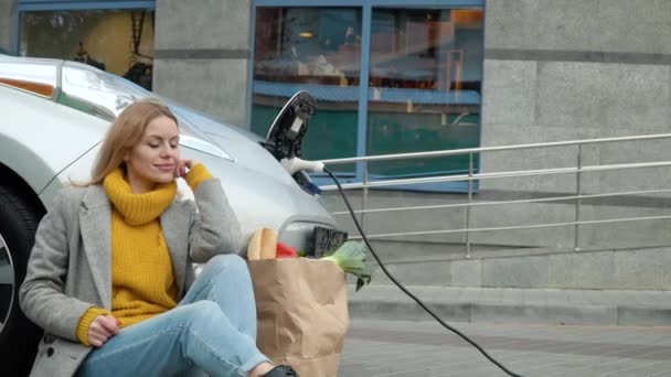 Een vrouw die naast haar elektrische auto zit. Elektrische auto opladen bij het elektrische benzinestation. Milieuvriendelijk vervoer — Stockvideo