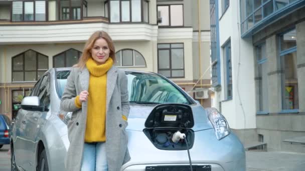 Een vrouw staat naast haar elektrische auto en kijkt naar de camera. Elektrische auto opladen bij het elektrische benzinestation — Stockvideo