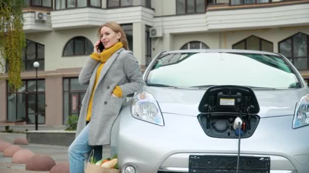 Γυναίκα μιλάει στο τηλέφωνο ενώ φορτίζει ένα ηλεκτρικό αυτοκίνητο — Αρχείο Βίντεο