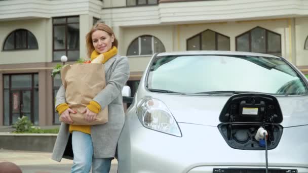 Een vrouw staat naast haar elektrische auto met boodschappen en kijkt naar de camera — Stockvideo