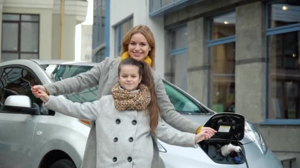 Una donna con sua figlia sta vicino alla sua auto elettrica e guarda la macchina fotografica. Caricare un'auto elettrica in una stazione di servizio — Video Stock