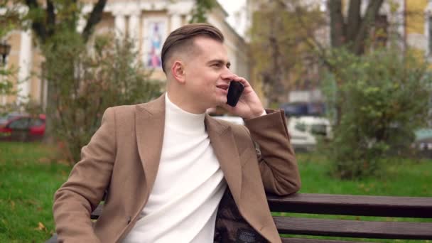 Элегантный улыбающийся мужчина разговаривает по телефону, сидя на скамейке на улице — стоковое видео