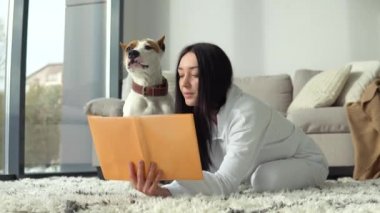 Genç bir kadın, köpeğiyle güzel, modern bir dairede kitap okuyor.