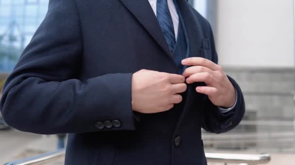 Close-up van een man die zijn jas dichtknoopt. Jonge elegante mannelijke zakenman in blauwe jas en jas in de buurt van kantoor centrum — Stockvideo