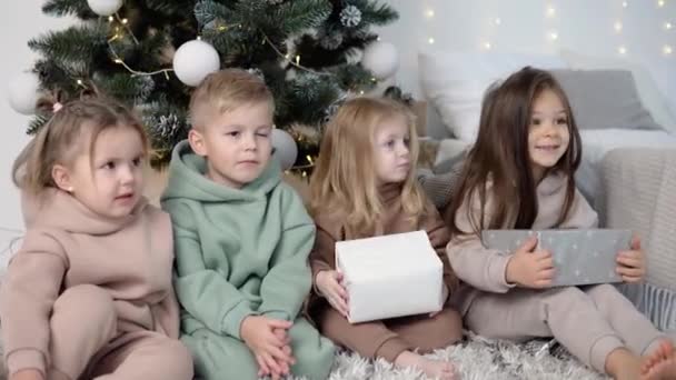 Çocuklar ellerinde hediyelerle Noel ağacının yanında oturuyorlar. — Stok video
