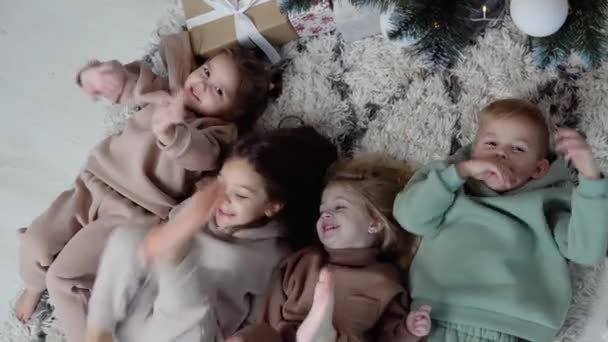 Quatro crianças doces deitam um tapete perto da árvore de Natal e acenam as pernas e as mãos. Vista de cima — Vídeo de Stock