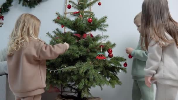 Süße Kinder in stylischen Freizeitanzügen schmücken einen Weihnachtsbaum — Stockvideo
