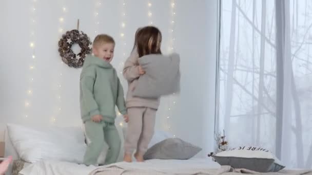 Niño y niña saltando sobre la cama con almohadas cerca de las decoraciones de Navidad — Vídeos de Stock