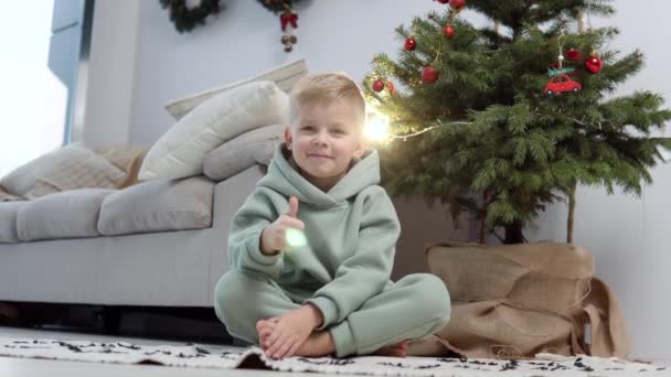 Blonďatý chlapec ve stylovém neformálním obleku sedí na koberci u vánočního stromku a ukazuje prstem super — Stock video
