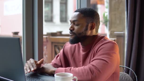 Afrykański amerykański młody człowiek komunikujący się przez telefon konferencyjny mówić patrząc na komputer w kawiarni — Wideo stockowe