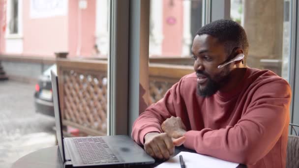 El joven afroamericano usa auriculares que se comunican por conferencia telefónica y habla mirando la computadora en la cafetería. Trabajo independiente desde la cafetería — Vídeo de stock