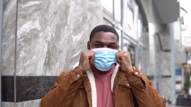 Tıbbi maskeli ve omuz çantalı siyah adam ofis merkezinin yanında duruyor. — Stok video