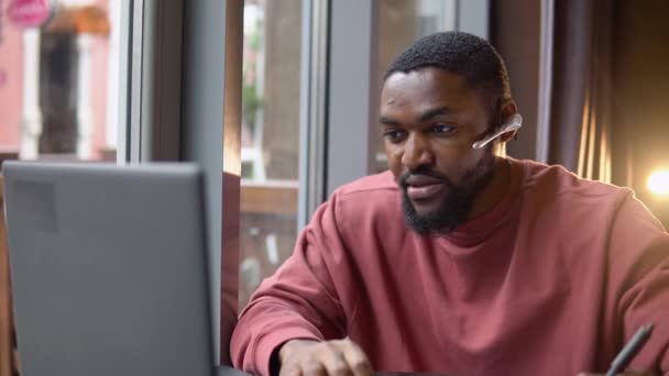 O jovem afro-americano usa auscultadores a comunicarem-se por teleconferência. Fala a olhar para o computador no café. Vídeo chat entrevista de emprego ou conceito de classe de curso de idiomas à distância — Vídeo de Stock