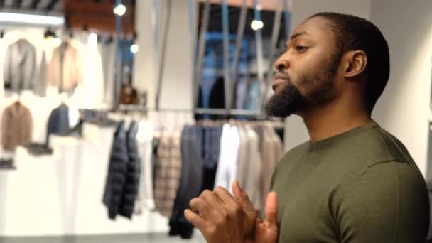 Een jonge, elegante Afro-Amerikaanse man in olijfgroen pak staat op een bank in een showroom of boetiek. — Stockvideo