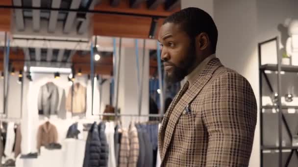 Joven hombre afroamericano confiado en la boutique de ropa de hombre de lujo eligiendo un nuevo traje. Tienda de ropa para hombre — Vídeo de stock