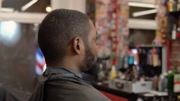 Un joven está sentado en una silla de peluquero frente a un espejo antes de un corte de pelo — Vídeo de stock