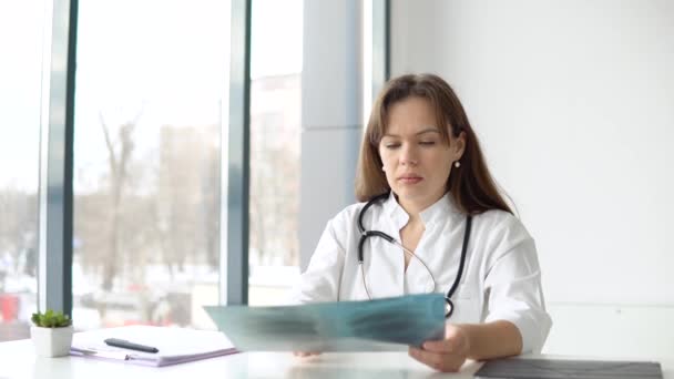 Una joven doctora caucásica que usa un abrigo médico blanco y un estetoscopio sostiene la radiografía y toma notas — Vídeo de stock