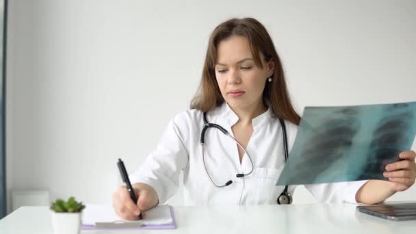 Jovem médica caucasiana vestindo casaco médico branco e estetoscópio escreve uma história médica em um caderno e segura o raio-x — Vídeo de Stock