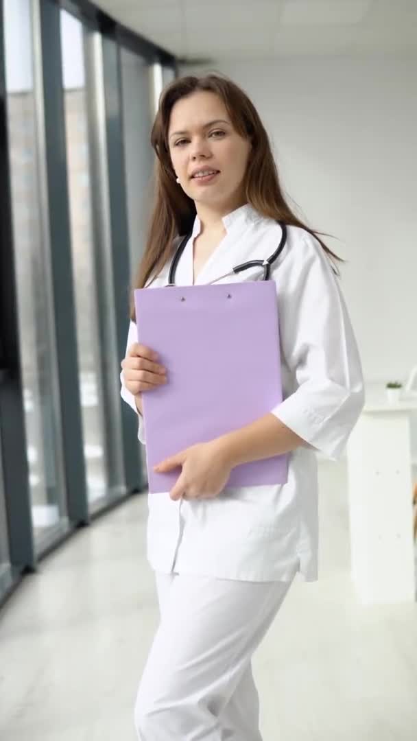 Gelukkige jonge blanke vrouw arts draagt witte medische jas en stethoscoop met documenten in haar handen kijken naar camera. Lachende vrouwelijke arts of verpleegkundige in het ziekenhuis kantoor. Verticale video — Stockvideo