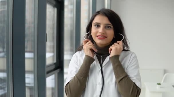 Happy young indian woman doctor in white medical coat wears a stethoscope and looking at camera. Smiling female physician posing in hospital office — Stock Video