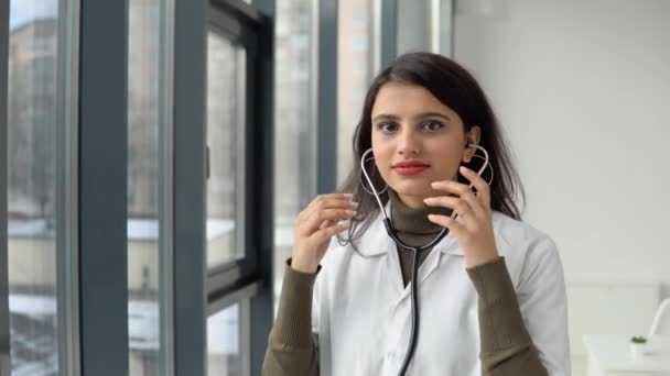 Szczęśliwy młody indyjski student medycyny ubrany w biały płaszcz medyczny i stetoskop patrzący w kamerę. Uśmiechnięta lekarka pozująca w gabinecie szpitalnym — Wideo stockowe
