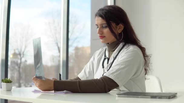 Jovem estudante de medicina indiana na clínica na prática. Médica de jaleco branco com estetoscópio examina raio-X e faz anotações — Vídeo de Stock