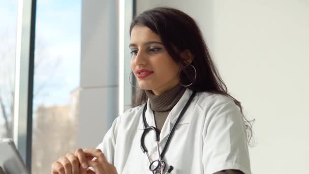 Индийская женщина-врач в белом халате со стетоскопом проводит онлайн-консультацию в своем кабинете — стоковое видео