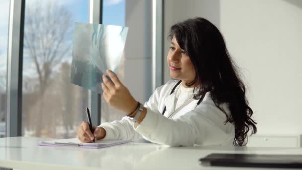 Jeune étudiant indien en médecine à la clinique en pratique. Femme médecin en manteau blanc avec stéthoscope examine les rayons X et prend des notes — Video